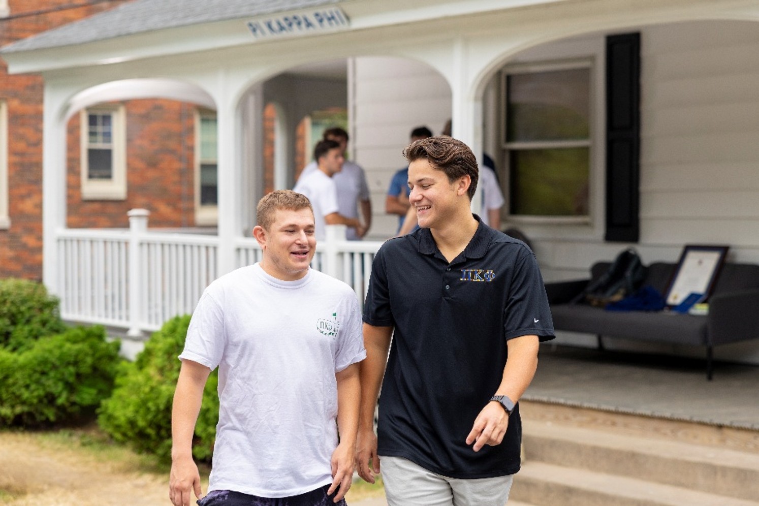 Two people laugh while walking in front of the Pi Kappa Phi house porch, with other students visible in the background.