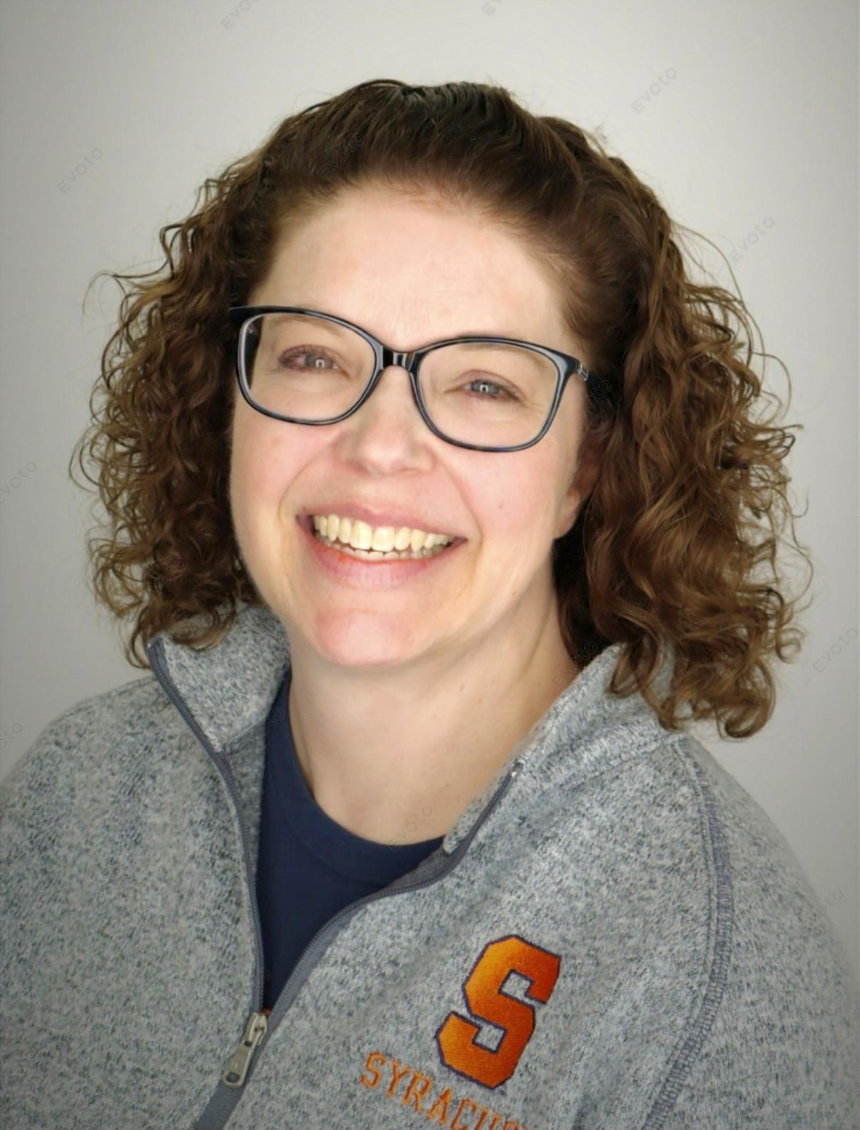 Professional headshot of a person with curly hair and glasses wearing a gray Syracuse quarter-zip pullover.