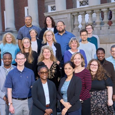 Group portrait of approximately 20 staff members standing on the steps outside a large brick building with columns.