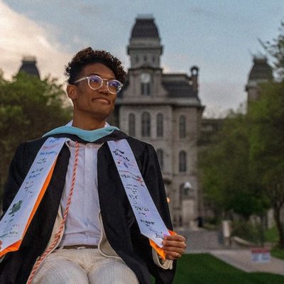 Graduate in cap and gown posing on a campus walkway, with a historic academic building in the background.