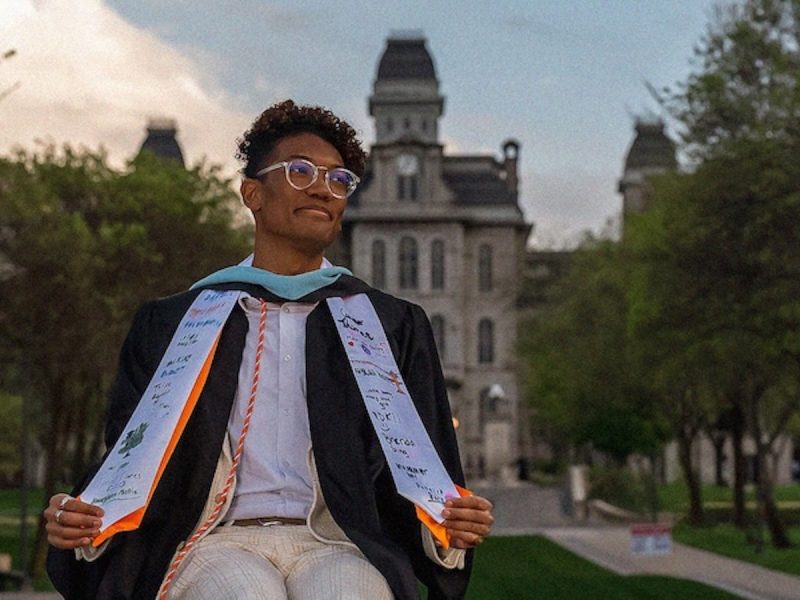 Graduate in cap and gown posing on a campus walkway, with a historic academic building in the background.