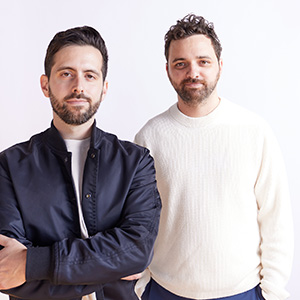 Two men standing together against a plain light background, one with arms crossed wearing a navy jacket, the other in a cream sweater, both with short beards.
