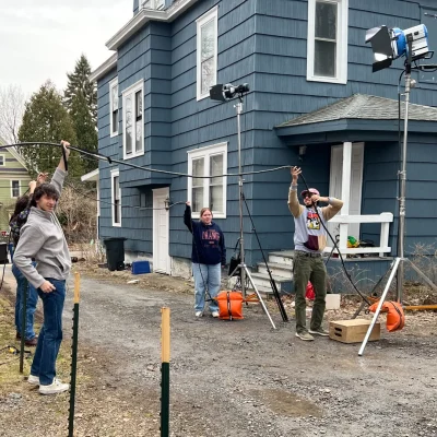 Students set up lighting equipment and cables outside a house during filming.