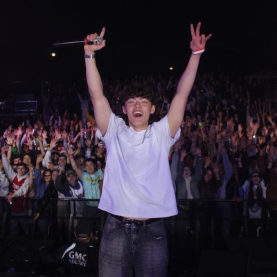A performer on stage holds a microphone aloft with one hand and flashes a peace sign with the other, arms raised in celebration. He's smiling broadly, wearing a white t-shirt and jeans, facing a large, enthusiastic crowd with hands raised at an outdoor nighttime concert.