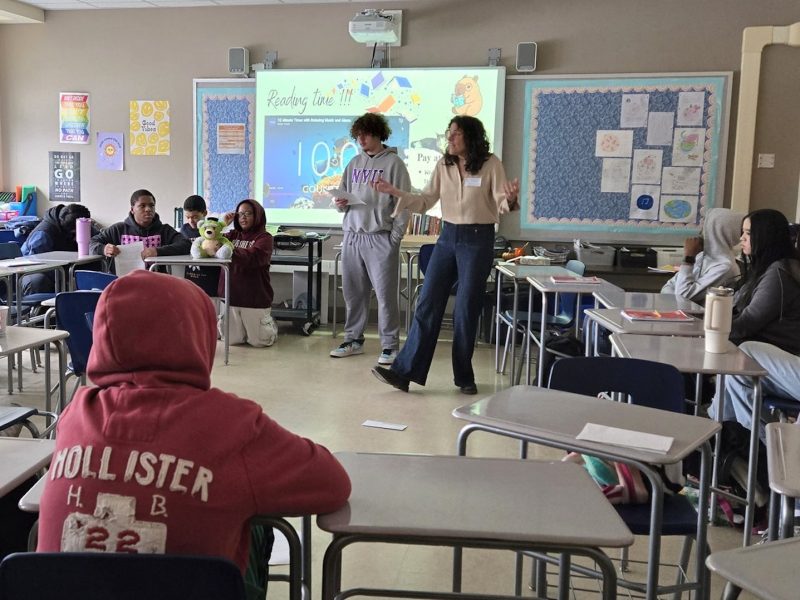 Classroom presentation in progress as students watch two speakers at the front of the room.