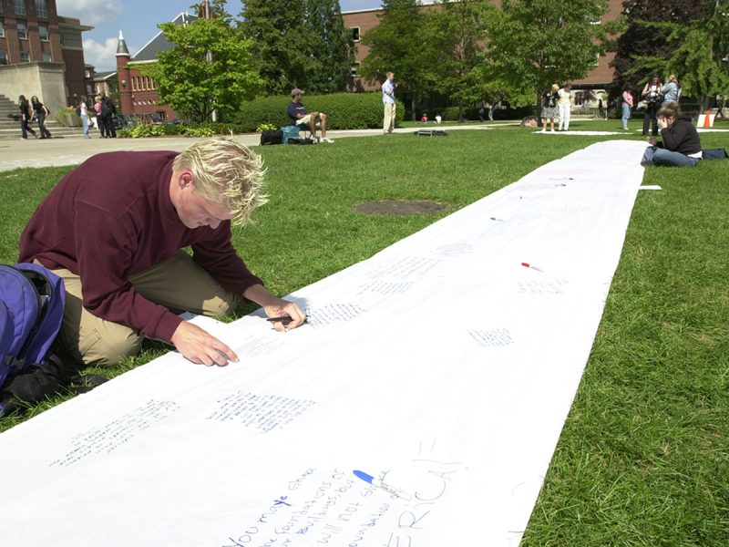 A young man in a maroon sweater kneels on grass writing a message on a long white fabric banner stretched across a lawn, with other people writing further down the banner and campus buildings visible in the background.