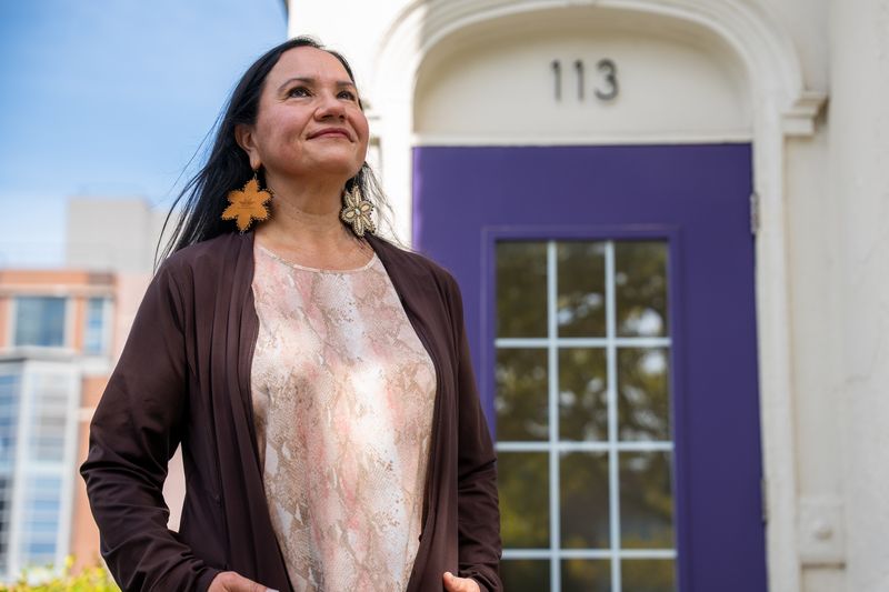 A woman wearing feather earrings stands outside a building with a purple door.