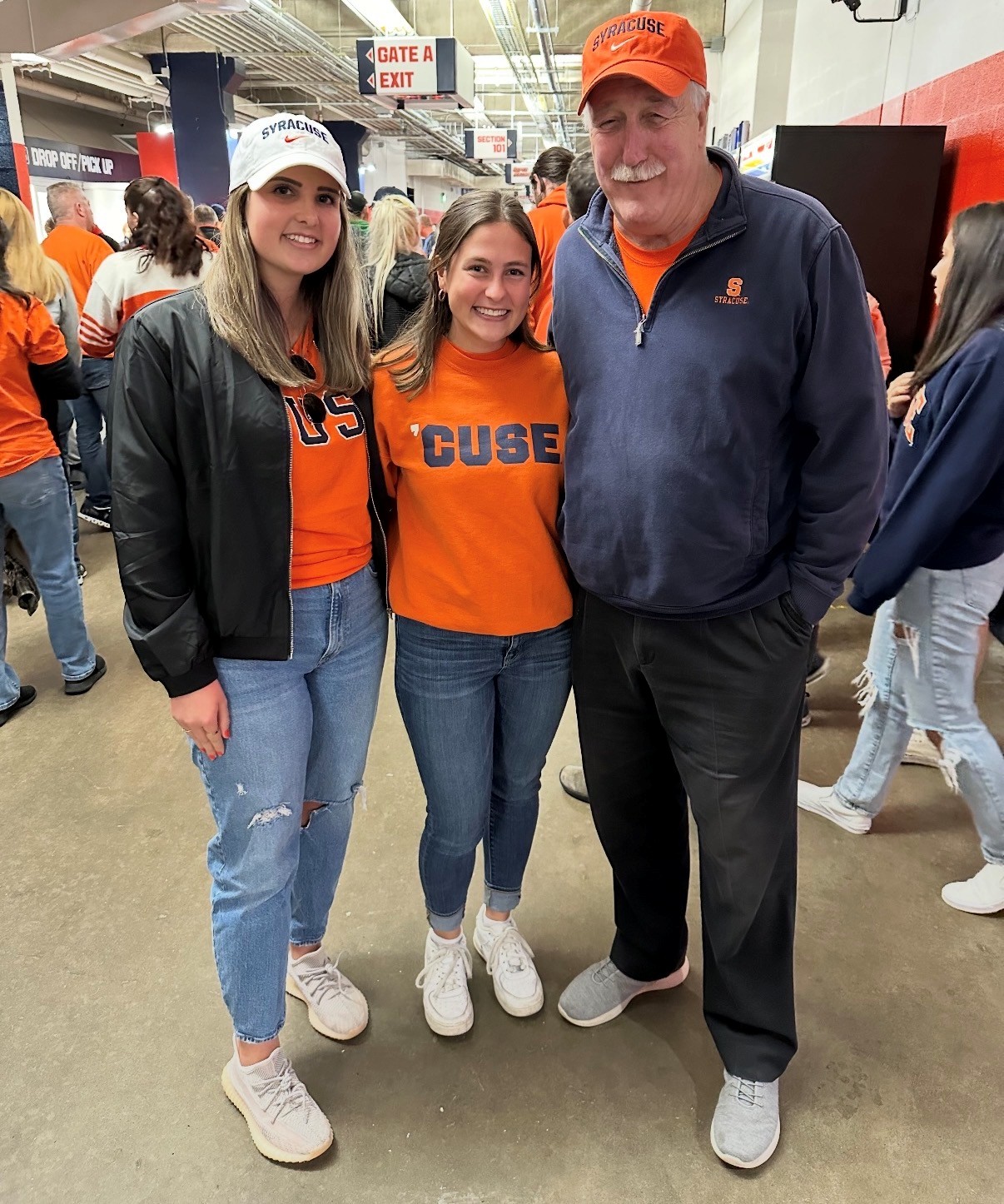 Three Syracuse fans in orange team apparel pose together inside a stadium concourse near Gate A.