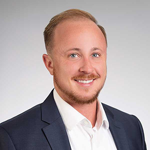 Headshot of a smiling man with reddish-blond hair and beard, in a navy suit, gray background.