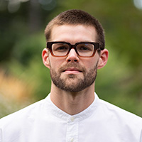 Headshot of a bearded man in glasses and a white shirt, against a blurred green outdoor background.