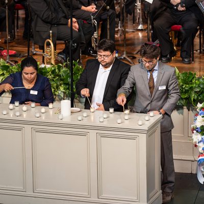 Three people light rows of votive candles at a memorial ceremony, with an orchestra and a patriotic floral wreath in the background.