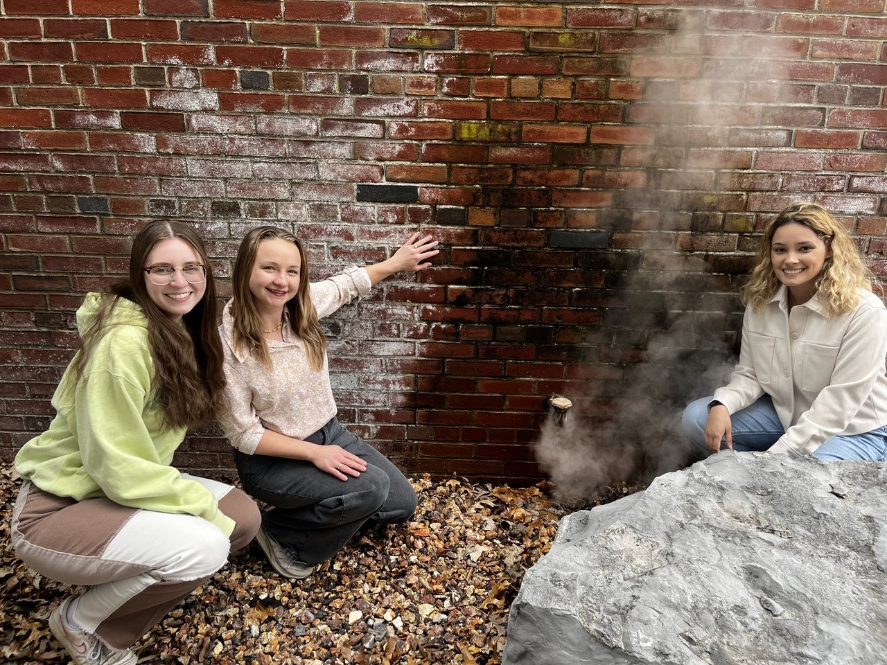 Three people kneeling beside a steaming ground vent near a brick wall.