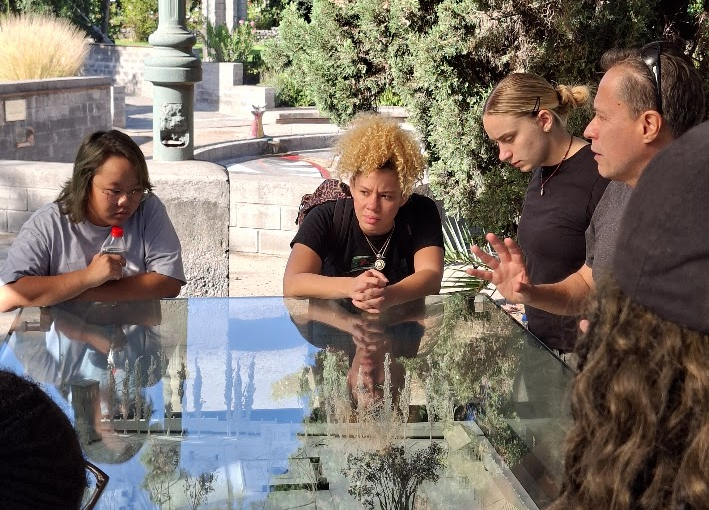 A small group leans over a glass-topped display case featuring a scale model of a garden or plaza, listening as a man gestures while explaining the model outdoors near a fountain and greenery.