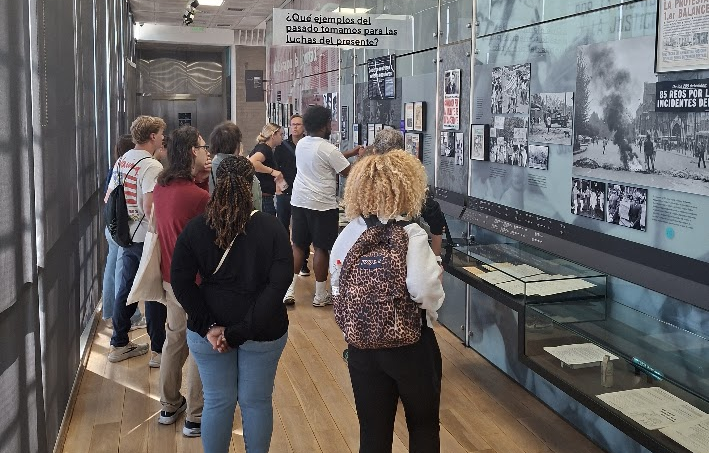 A group of visitors views a museum exhibit lining a glass-walled corridor, featuring historical black-and-white photographs and newspaper clippings with a Spanish-language question displayed on a sign overhead.