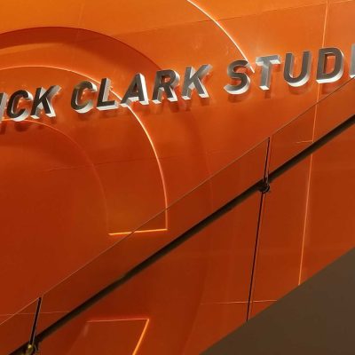 Sign reading “Dick Clark Studios” mounted on a bright orange interior wall above a modern staircase with glass railing.