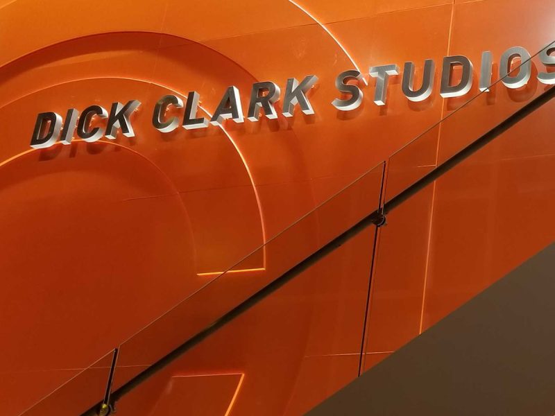 Sign reading “Dick Clark Studios” mounted on a bright orange interior wall above a modern staircase with glass railing.