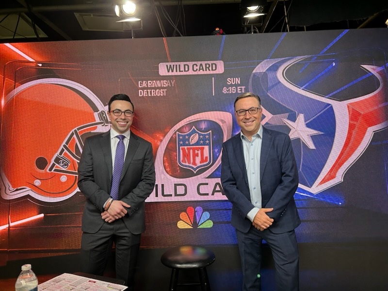 Two people in suits stand smiling in front of an NFL Wild Card game backdrop