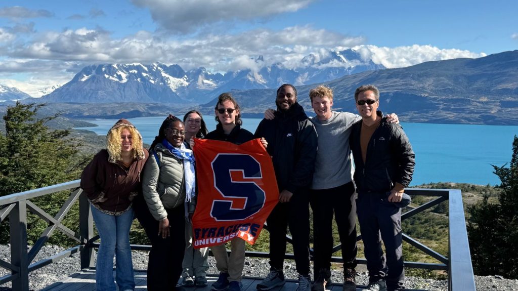 A group poses on an overlook platform in Patagonia, holding an orange Syracuse University flag, with snow-capped mountains and a turquoise lake stretching behind them.