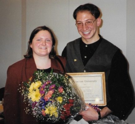 A person with a bouquet stands beside a person holding a framed Syracuse University certificate.