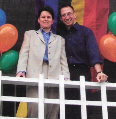 Two smiling people lean on a white railing in front of a rainbow flag and colorful balloons.