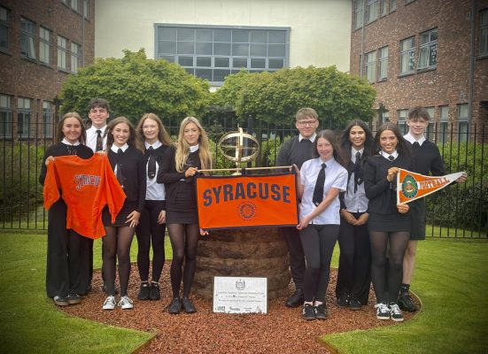 Ten Lockerbie Academy students in school uniforms hold Syracuse University banners and a sweatshirt beside the Lockerbie Academy–Syracuse University Cairn.