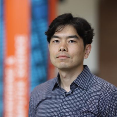 Professional headshot of a person in a patterned button-down shirt standing indoors, with a softly blurred background featuring orange and blue accents.