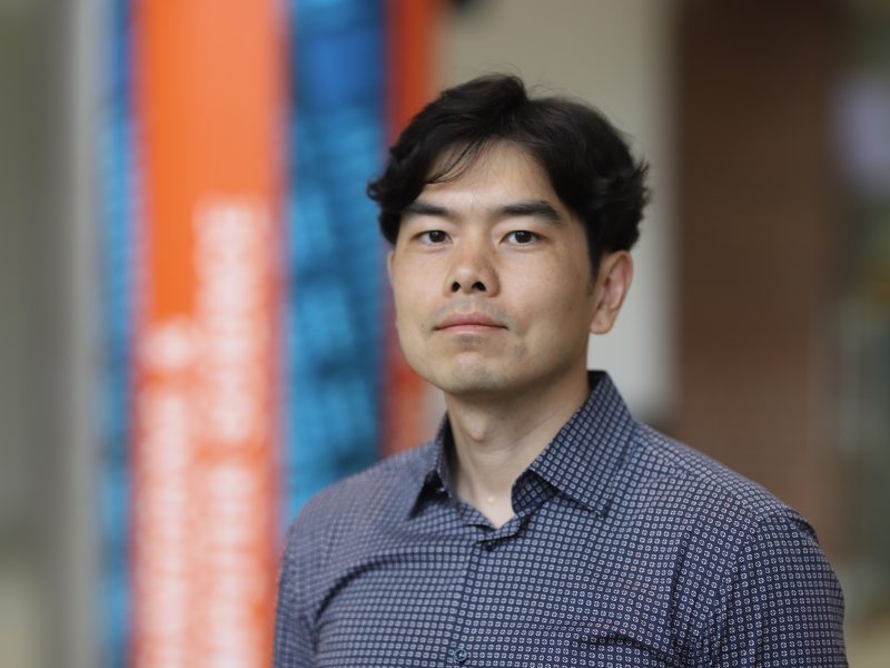 Professional headshot of a person in a patterned button-down shirt standing indoors, with a softly blurred background featuring orange and blue accents.