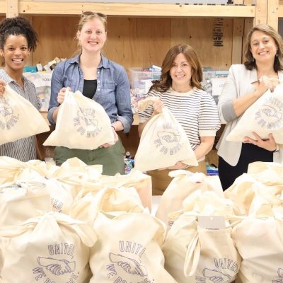 Six volunteers stand behind a table filled with donation bags, holding canvas tote bags in a community supply-packing workspace.