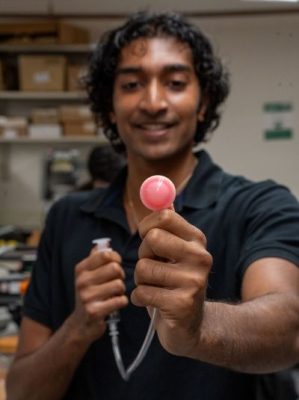 Ruben John smiles while holding up a small pink gripper device attached to a syringe-like mechanism in a lab setting.