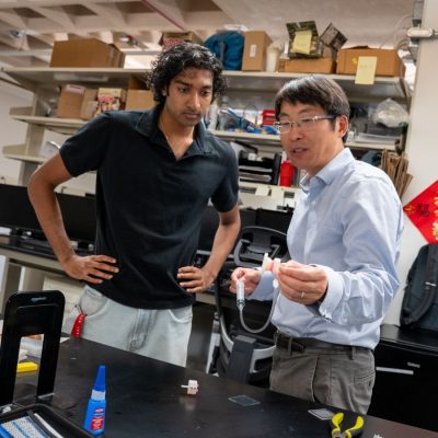 Student Ruben John, left, looks on as his professor, Wanliang Shan, explains a small gripper device he holds up at a lab workbench cluttered with tools and research materials.