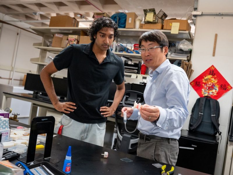 Student Ruben John, left, looks on as his professor, Wanliang Shan, explains a small gripper device he holds up at a lab workbench cluttered with tools and research materials.