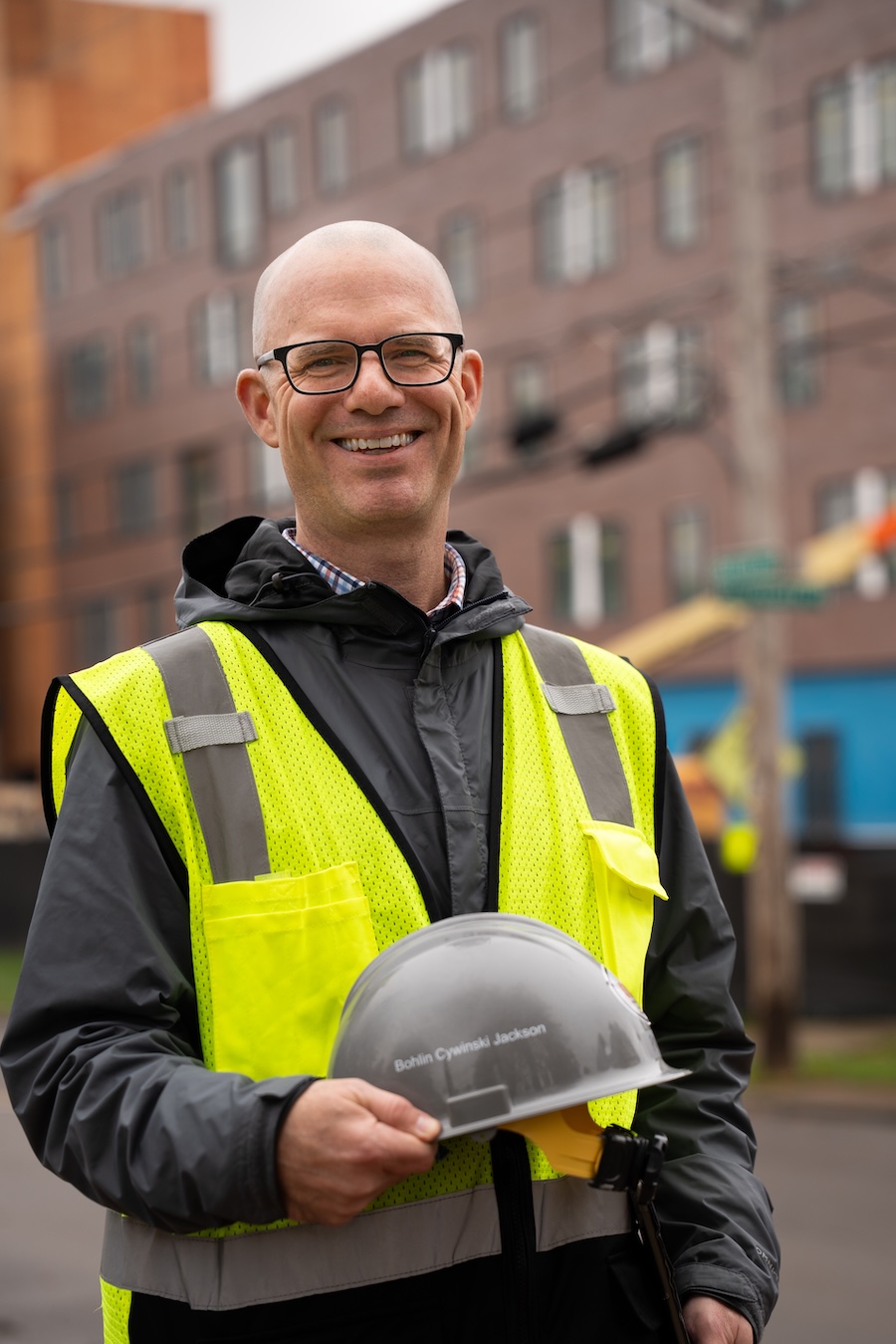 Person in a safety vest holds a hard hat in front of a residence hall construction site.