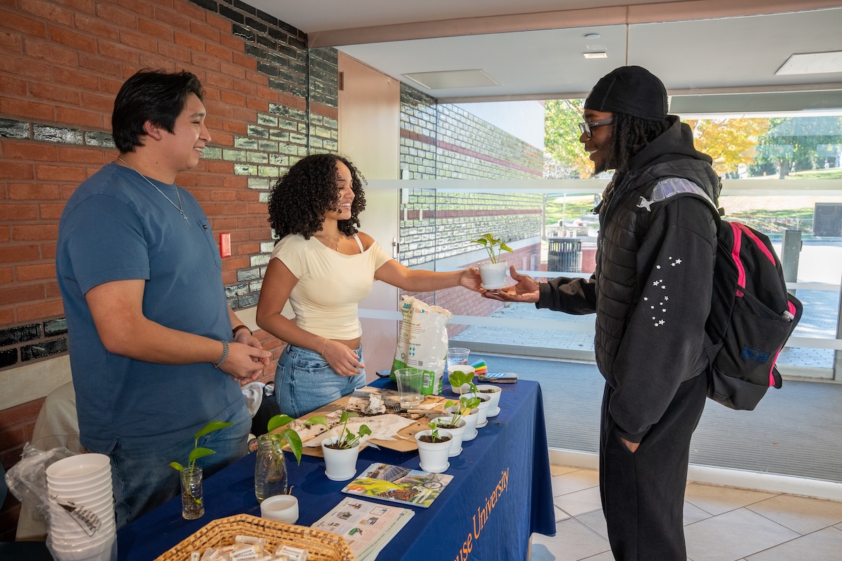 Two students at a campus table hand a small potted plant to a smiling passerby, with propagation jars and more potted cuttings on the table.