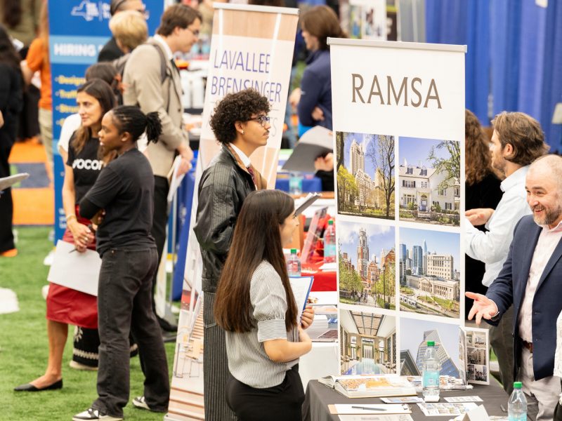 A recruiter at a booth smiles while talking with a student at a crowded career fair.