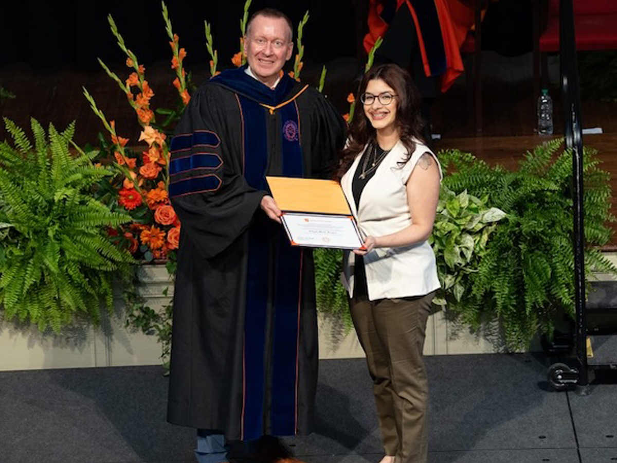 A woman in a white vest holds a certificate on stage beside a man in academic regalia.