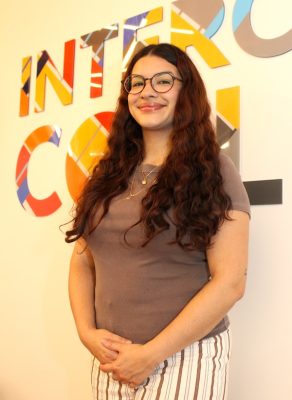 A smiling person stands in front of a colorful wall sign.