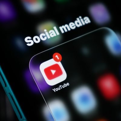 Close-up of a smartphone displaying the YouTube app icon with a notification badge, set against a blurred social media-themed background.