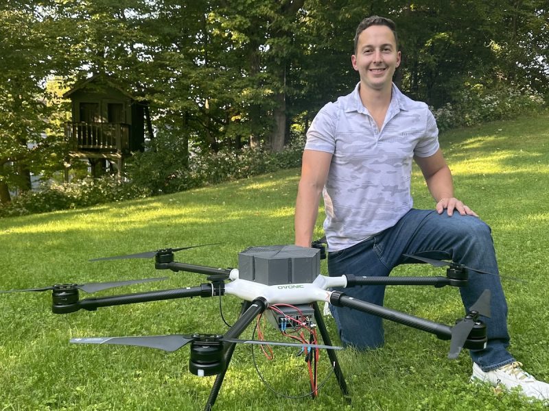 Person kneeling beside a large drone on a grassy lawn with trees and a small cabin in the background.