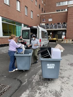 Three people unload a white van into two large gray rolling bins outside a brick building, collecting donated items like blankets, a laundry basket, and furniture.