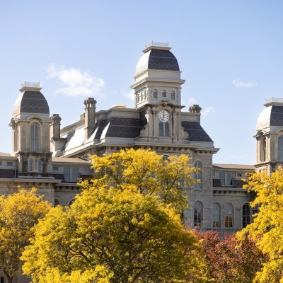 Historic stone academic building with a central clock tower and three domed turrets rising above bright yellow autumn trees under a clear blue sky.