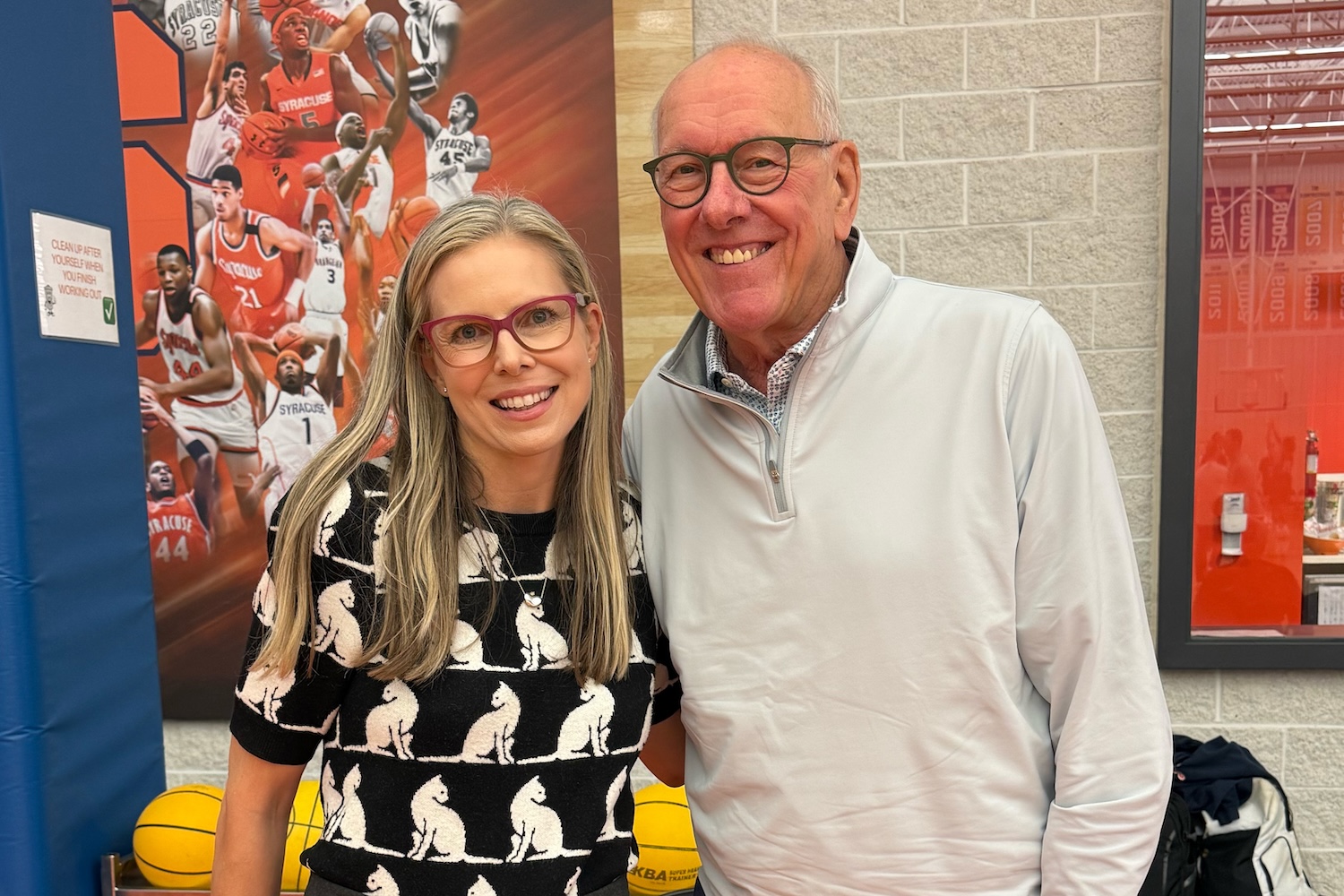 Two people standing together in a gymnasium with Syracuse basketball imagery in the background.