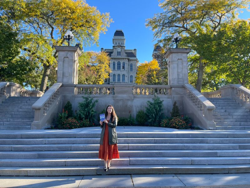 A woman in an orange dress and green jacket holds up a book on the steps below Hall of Languages.