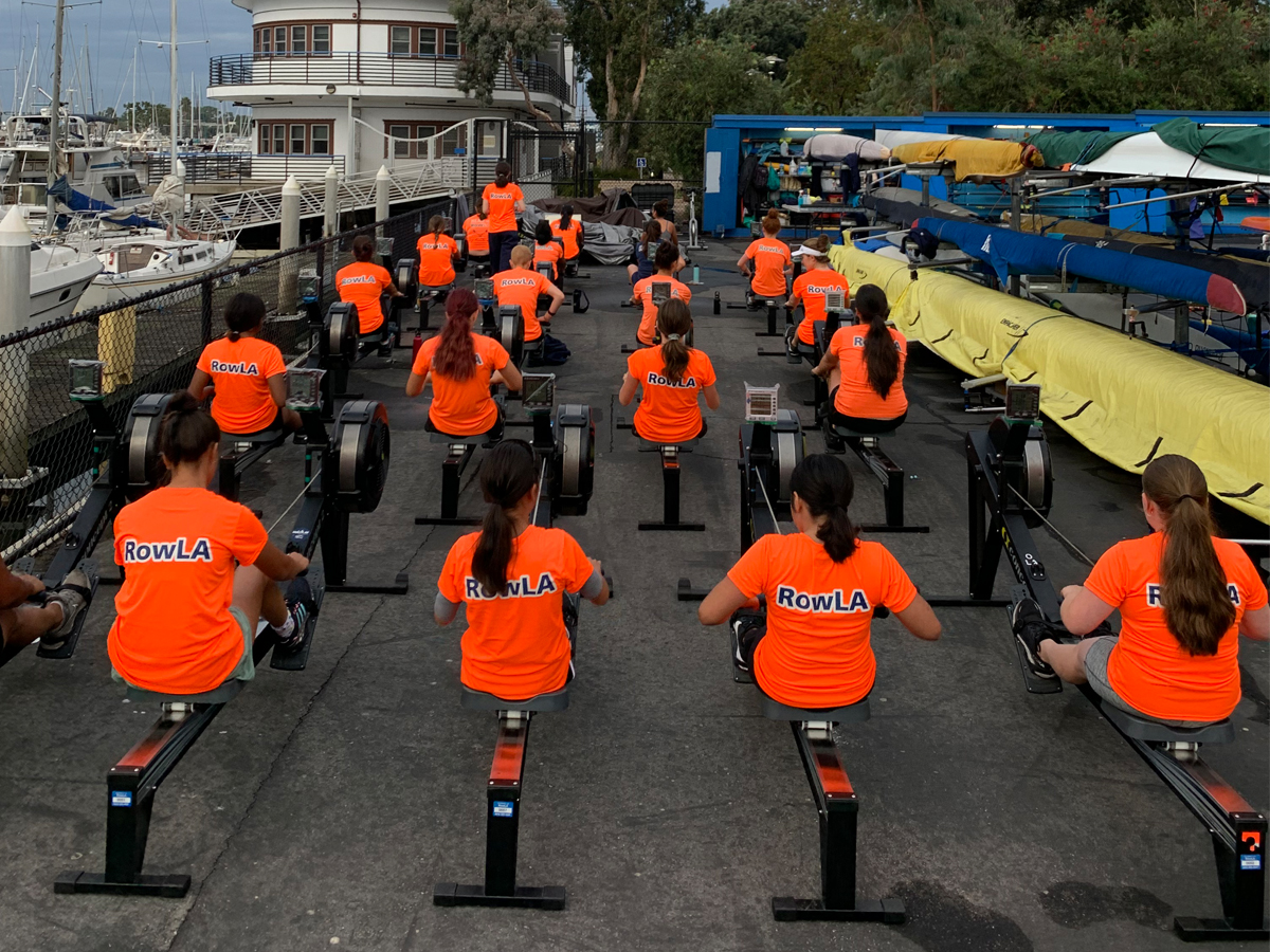Rowers in orange RowLA shirts train on indoor rowing machines at a marina dock