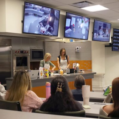 Two women cook in a demonstration kitchen as monitors and a caption screen display the lecture.