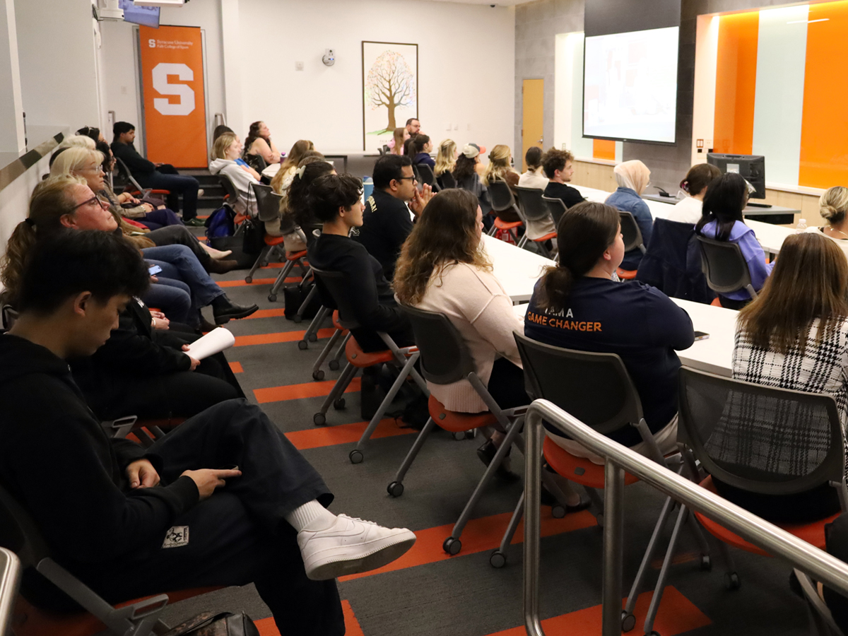 Audience members seated in rows watch a presentation on a screen in a classroom.