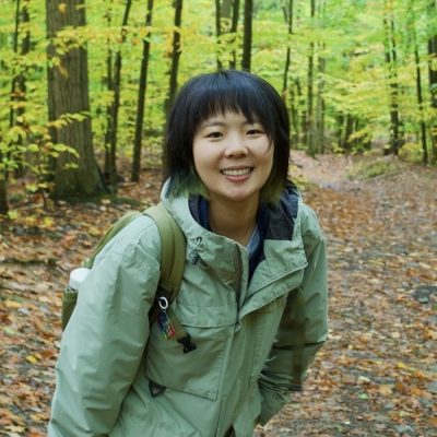 Person with a backpack and light jacket standing on a leaf-covered forest trail surrounded by autumn trees.