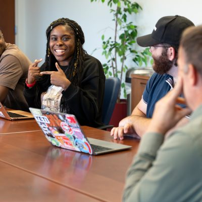 A smiling student points at herself while classmates and an instructor listen at a table.