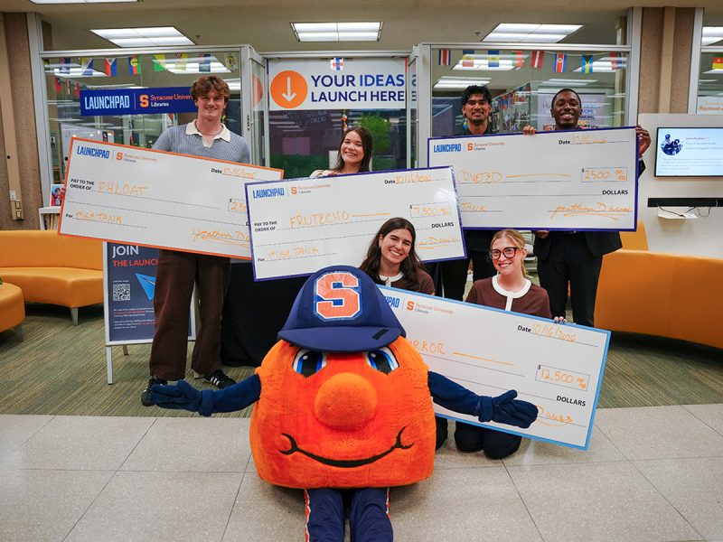 Otto the Orange poses with ’Cuse Tank winners holding oversized prize checks in Bird Library.