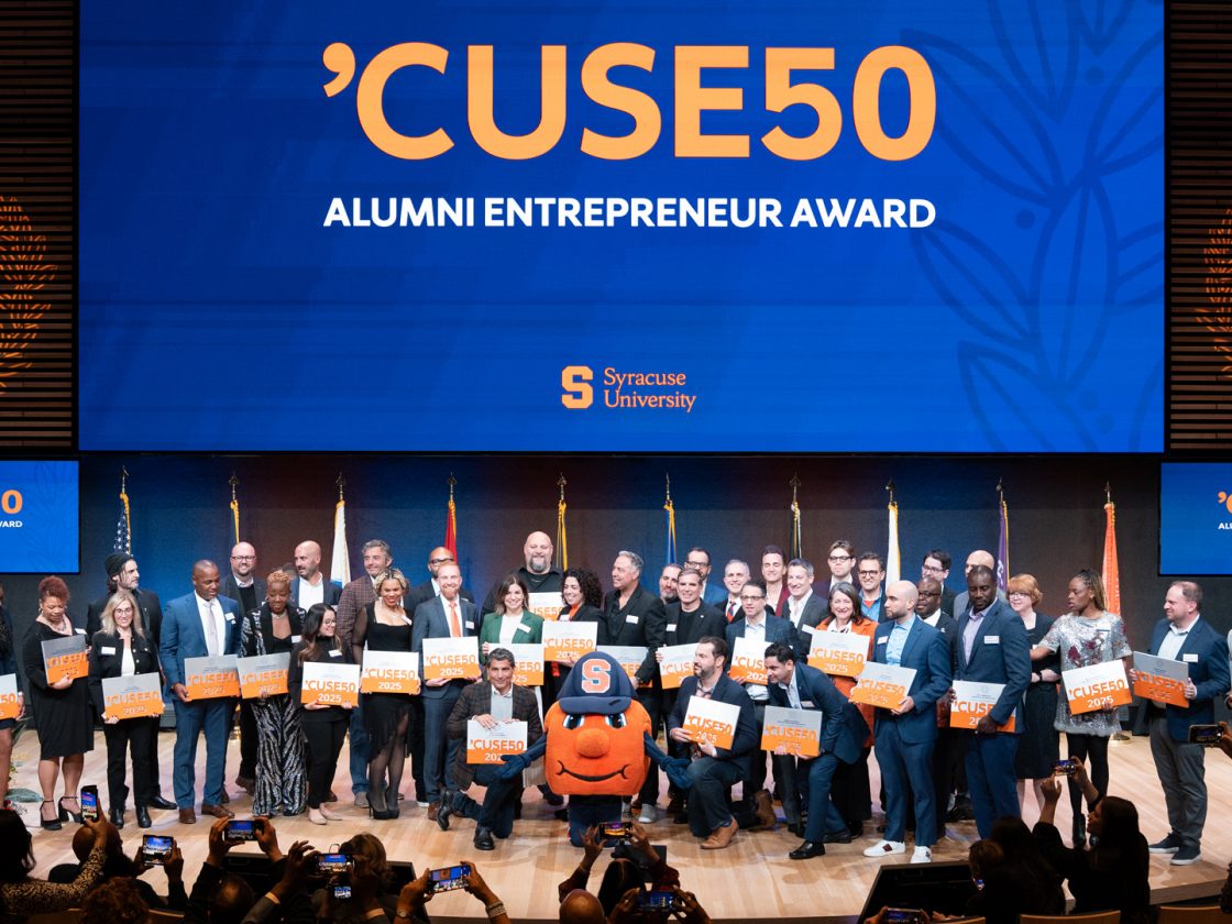 Dozens of ‘CUSE50 honorees hold 2025 plaques on stage with Otto beneath a large ‘CUSE50 screen.
