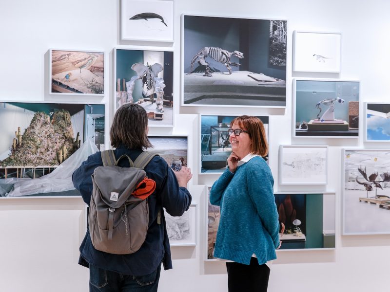 Visitors view and discuss a gallery wall of framed photographs featuring museum exhibits, fossils, and natural history displays.